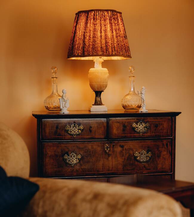 A glowing lamp on a Georgian chest of drawers beside an armchair in The Salwey Lodge